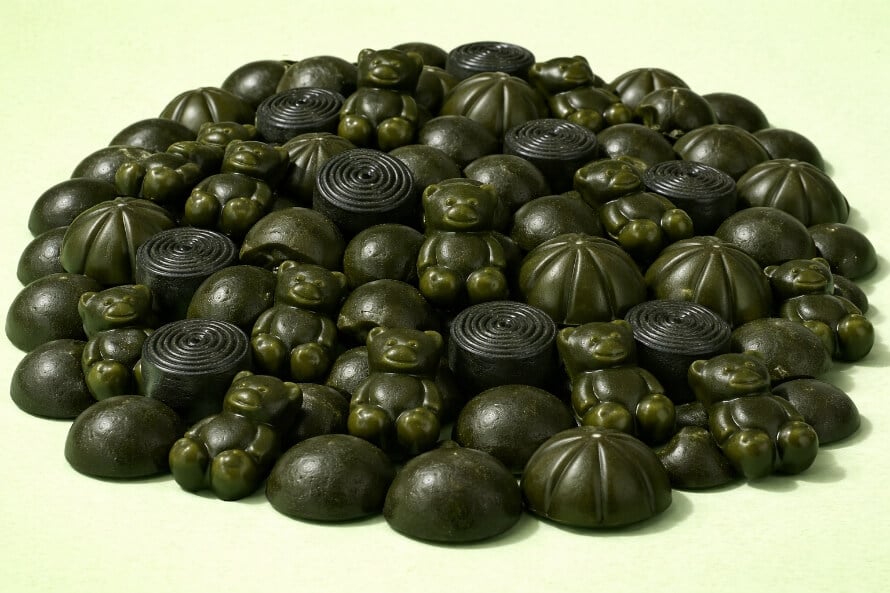 Large pile of assorted greens gummies in multiple shapes, including bears, spirals, and domes, on a light green background.